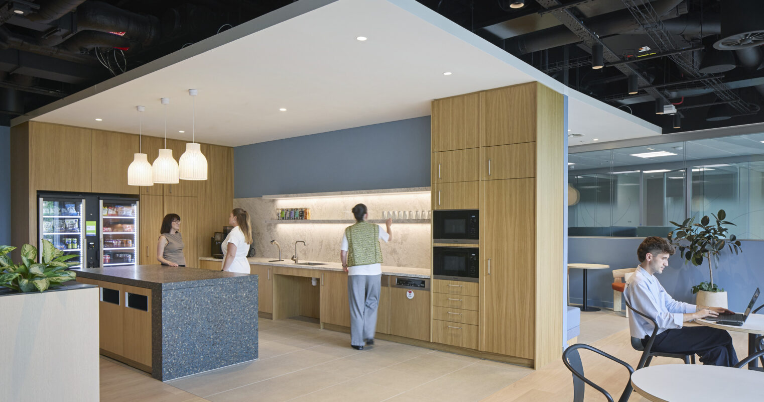 A modern office kitchen features warm wood cabinetry and sleek stone countertops. Three individuals engage at the kitchen island, while a fourth works at a nearby table. The design showcases efficient use of space, with integrated appliances and natural light enhancing the inviting atmosphere.
