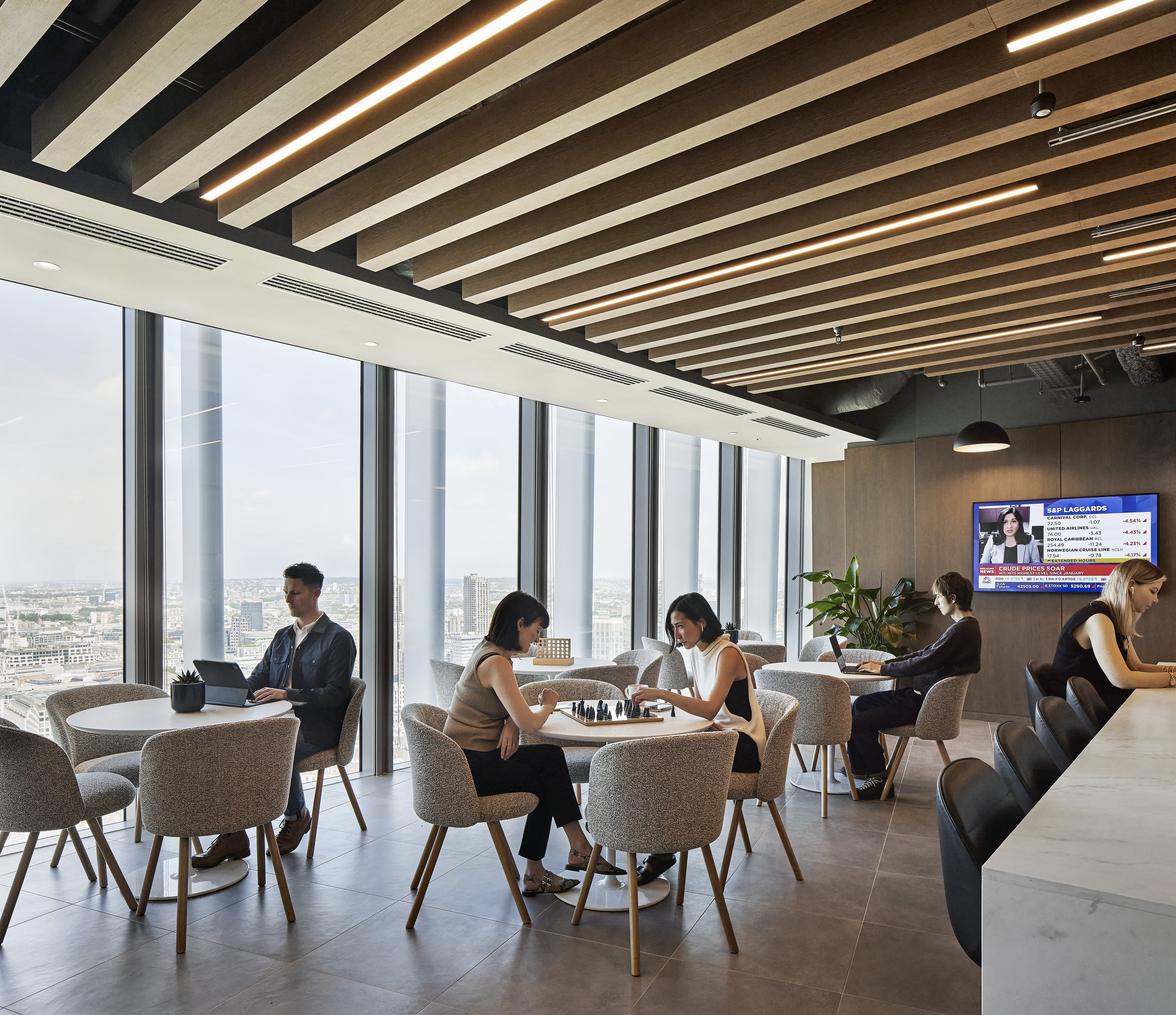 A modern office lounge featuring light-colored wooden beams on the ceiling, large glass windows offering expansive city views, and stylish furnishings. Comfortable armchairs and small round tables foster collaboration, while individuals engage in discussions or work. A large screen displays news updates, enhancing the dynamic environment.