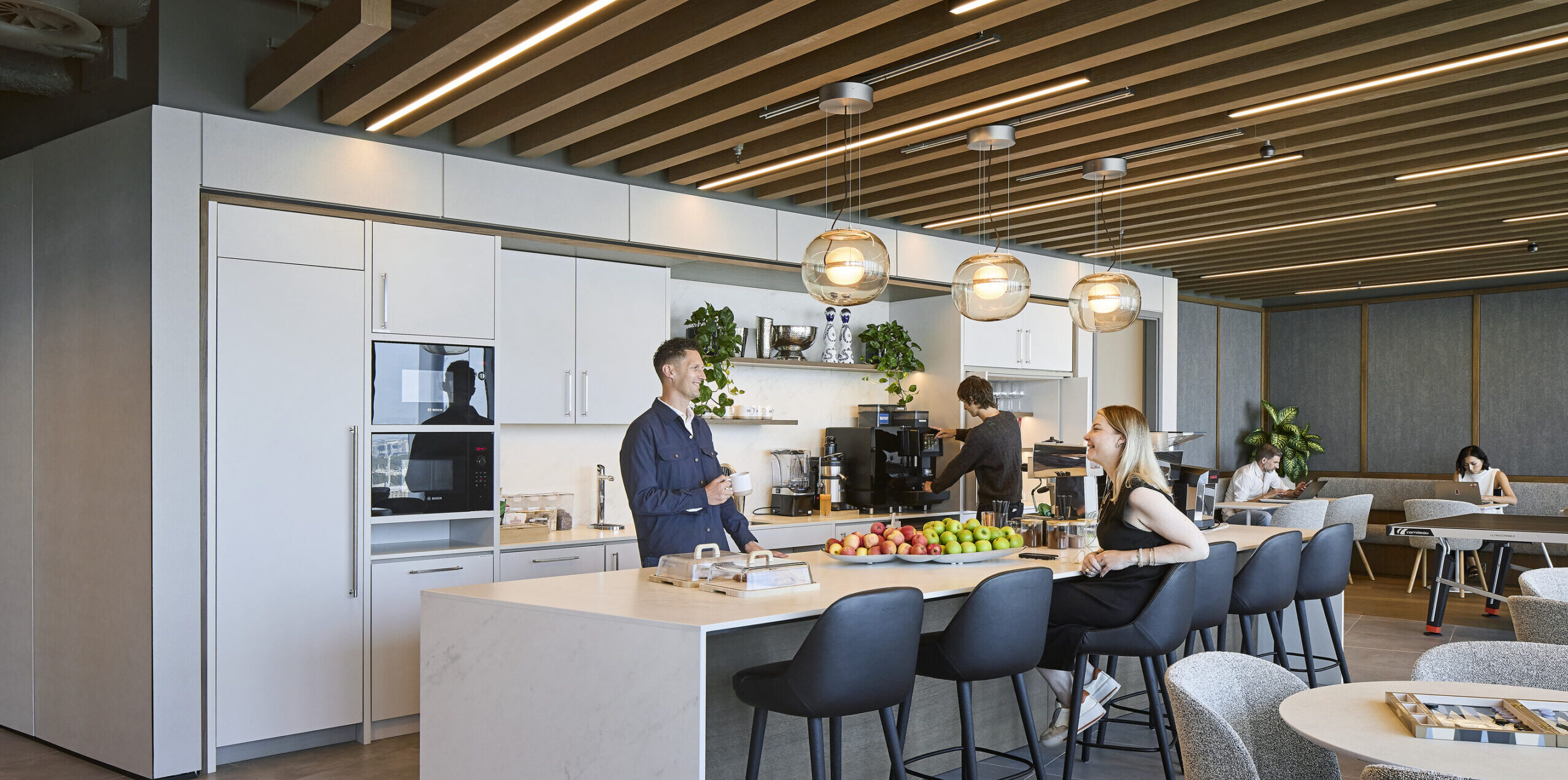A modern office kitchen featuring sleek, white cabinetry and quartz countertops. Pendant lights illuminate the workspace, enhancing the warm wooden ceiling beams. Individuals interact casually at a spacious island, surrounded by stylish bar stools and fruit displays, emphasizing a contemporary yet inviting atmosphere for collaboration and relaxation.