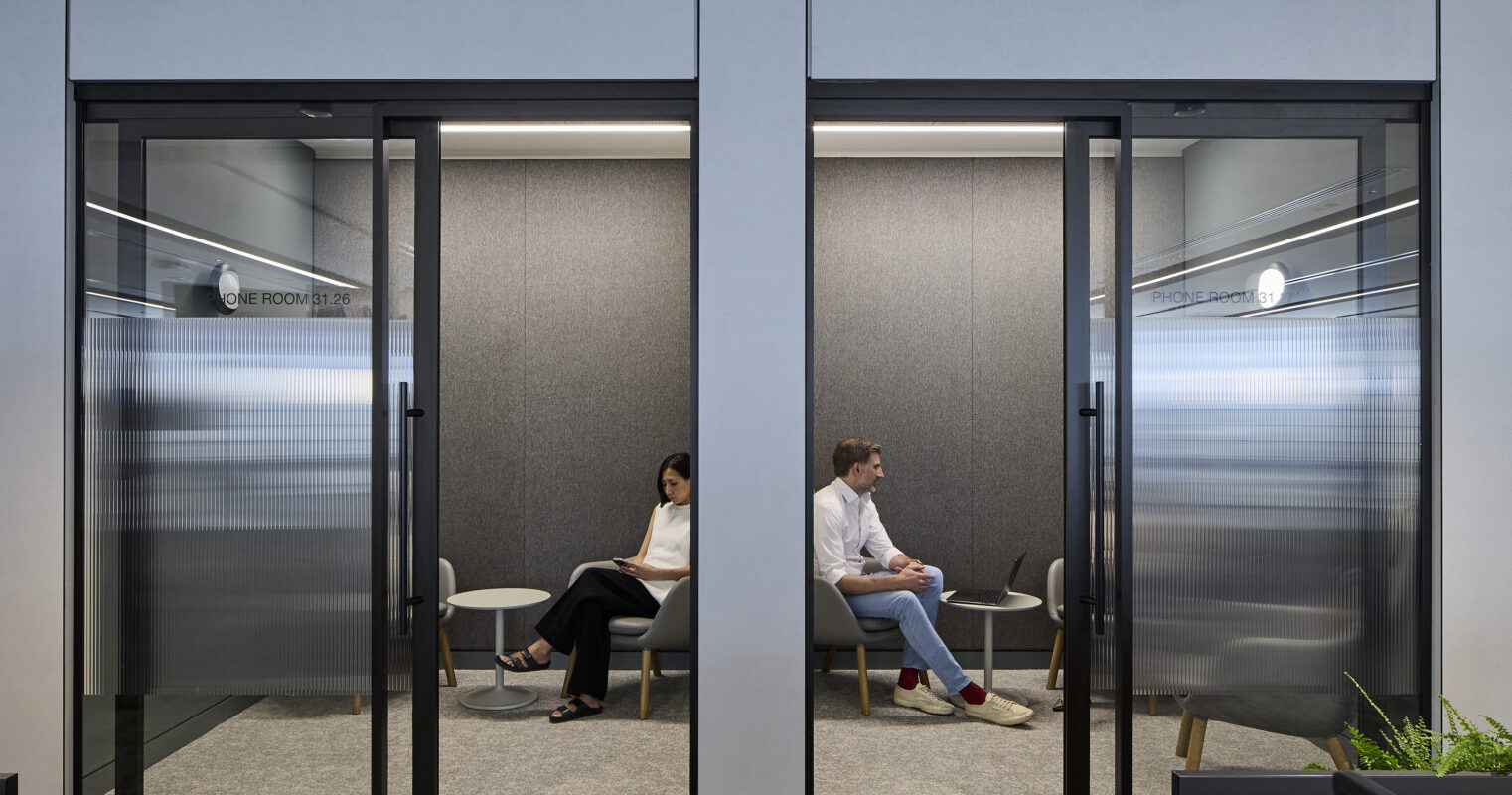 Two modern meeting pods are separated by a glass wall in an office environment. Each pod features sleek, minimalist furniture, including a small round table and comfortable seating. Soft gray acoustical panels enhance sound absorption, while natural light filters through the transparent surfaces, promoting a productive atmosphere.