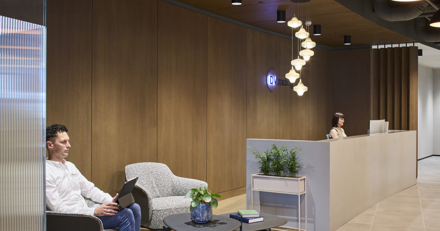 A modern office reception area featuring warm wood paneling and soft, ambient lighting. A man sits comfortably in a stylish armchair, working on a laptop. In the background, a minimalist reception desk displays greenery and decorative elements, creating an inviting and professional atmosphere.