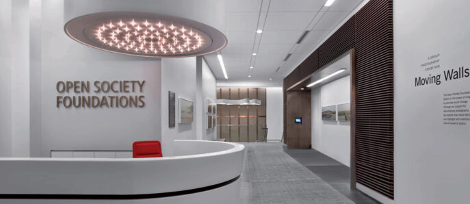 Modern minimalist reception area featuring a sleek white circular desk with a radiant red accent, cylindrical overhead lighting installation, and clean lines contrasting with the soft gray floor and geometric ceiling patterns. Contemporary art enhances the serene ambience.