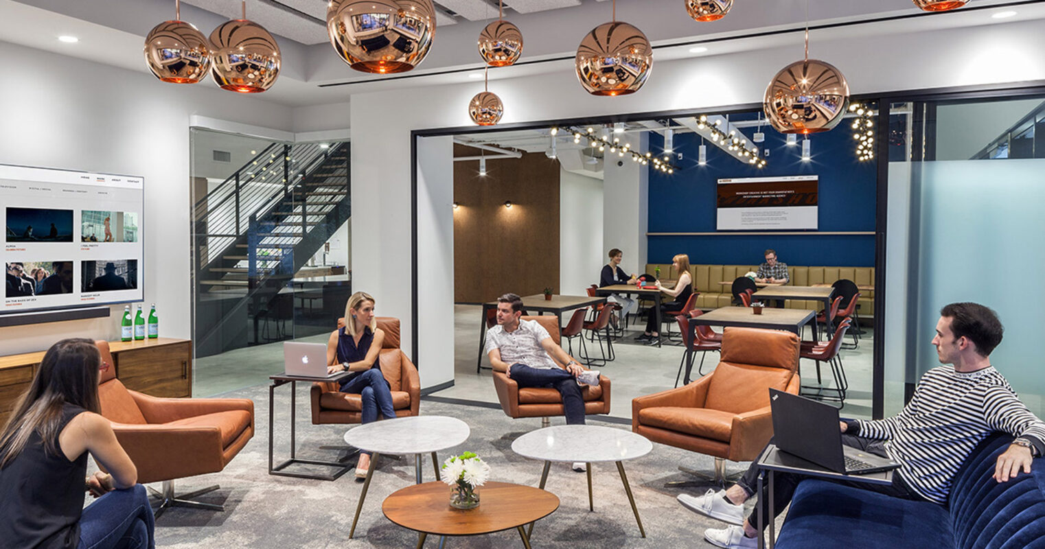 Modern open-plan office space featuring eclectic mix of traditional and contemporary furniture, copper pendant lights, and a two-tiered mezzanine for additional seating areas, fostering a vibrant collaborative atmosphere.