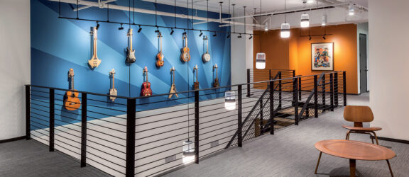 Modern music studio lobby with a collection of hanging guitars as a focal point. The space features contrasting blue and orange walls, industrial-style lighting, and minimalist furniture that complements the clean, creative environment.