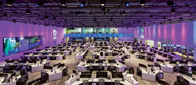 Spacious modern office featuring rows of white workstations with ergonomic chairs, ambient pink and purple lighting, and a high ceiling with grid lighting enhancing the contemporary atmosphere.