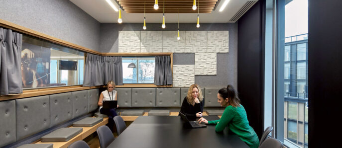 Modern corporate meeting room featuring a long, dark wooden table, with overhead yellow pendant lighting. Acoustic wood slat ceiling complements the gray upholstered wall panels. Two individuals engage in conversation, highlighting the space's collaborative atmosphere.