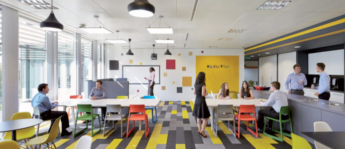 Modern office break room with vibrant yellow accents and geometric patterns. Natural light filters through large windows, illuminating the eclectic mix of seating options and interactive, collaborative spaces.