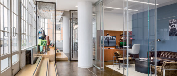 Modern office foyer featuring glass partitions, a polished concrete floor, and a white structural column. The space combines industrial elements with contemporary design, including a sleek reception desk and comfortable lounge seating by a large window with abundant natural light.