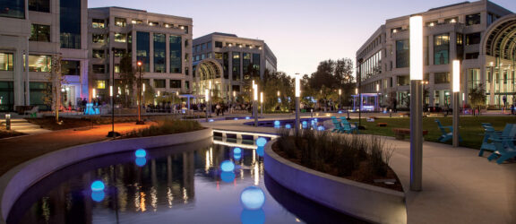 Modern urban plaza at dusk with symmetrically arranged buildings flanking a serene water feature, accented by illuminated blue spheres, and landscaped pathways leading to a central green space with benches for relaxation.