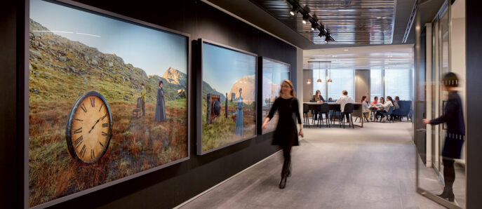 Modern office hallway featuring oversized wall clocks seamlessly integrated into pastoral landscape art, with recessed lighting and a distant collaborative work area.
