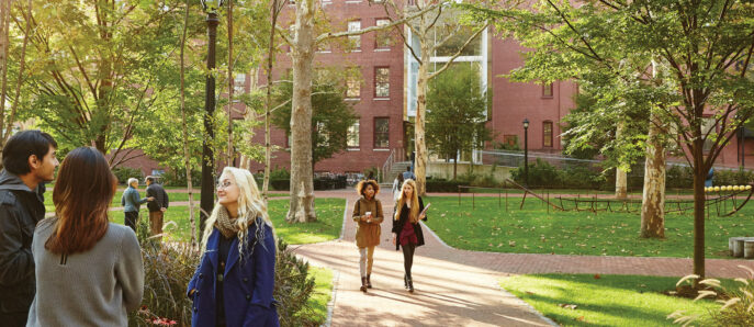 Individuals walking through a harmoniously designed landscape with lush greenery, framed by traditional red brick buildings, embodying a classic campus ambiance with thoughtful integration of nature and architecture.