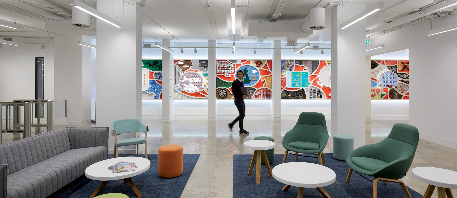 Bright, open-plan office lobby featuring a minimalist aesthetic with a variety of seating options, including teal armchairs, a grey sofa, and white round tables. Exposed ceiling with industrial lighting complements colorful, abstract wall art, creating a vibrant, welcoming space.