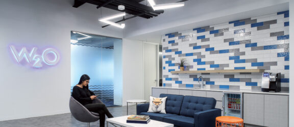 Modern office lounge featuring a blue sofa with geometric cushions, a round glass-top coffee table, and a wingback chair. The wall is accented with blue and white tiles, complemented by a backlit company logo. The setting is enhanced by sleek black exposed ceiling beams.