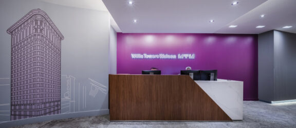 Elegant office lobby featuring a vibrant fuchsia accent wall with Willis Towers Watson branding and a line drawing of a skyscraper, complemented by a sleek, wooden reception desk and modern, recessed ceiling lighting.