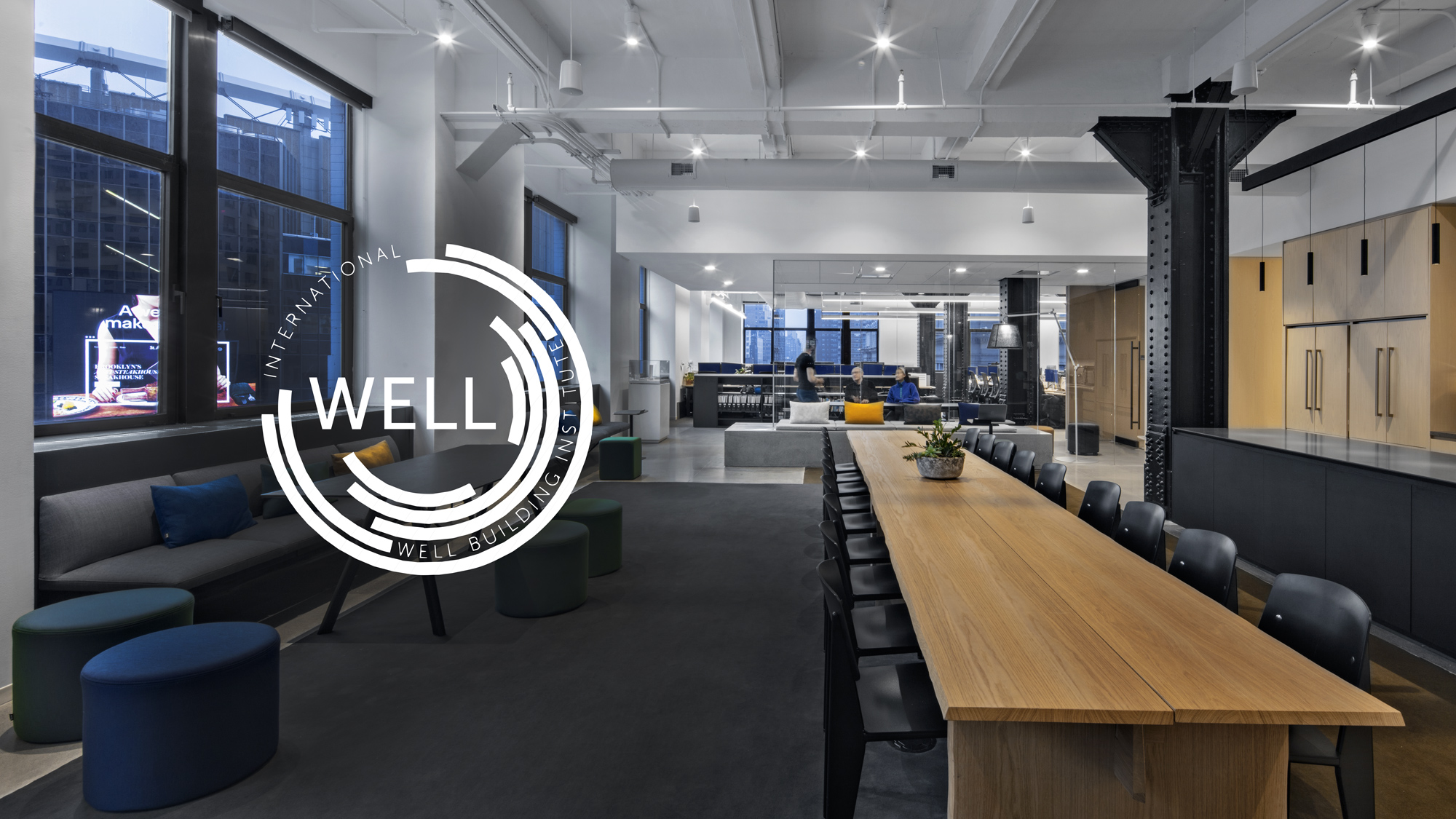 A modern office interior with industrial design elements, a large wooden table, black chairs, and a seating area with a logo of the International WELL Building Institute overlaid on the image.