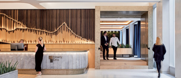 Modern office lobby with a curved wooden reception desk. Marble flooring transitions to a wooden passageway framed by elegant vertical wooden slats. Ambient lighting accentuates the smooth, natural materials and open, welcoming space.