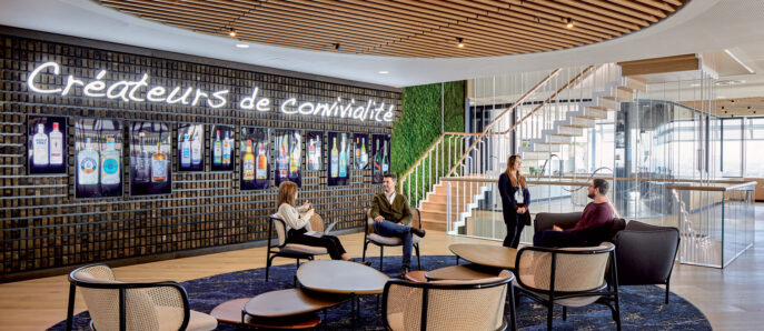 Contemporary office lounge with wooden slatted ceiling curving downward, incorporating ambient lighting. Dark marble floor hosts a cluster of elegant rattan chairs on a circular navy rug. A living green wall and illuminated brand motto enhance communal aesthetic. People converse and work on laptops, suggesting dynamic use.