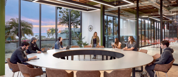 Modern office space with a central oval table surrounded by tan leather chairs. Employees engage in discussion, a dog rests nearby. Glass partitions and an exposed ceiling with hanging linear lights create an open and collaborative environment. Coastal vista visible through floor-to-ceiling windows.