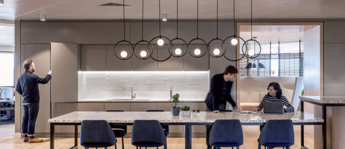 Modern kitchen and dining space with integrated lighting, featuring a marble island, dark upholstered chairs, and pendant fixtures. A man observes the sleek cabinetry, while two individuals collaborate at the dining table.