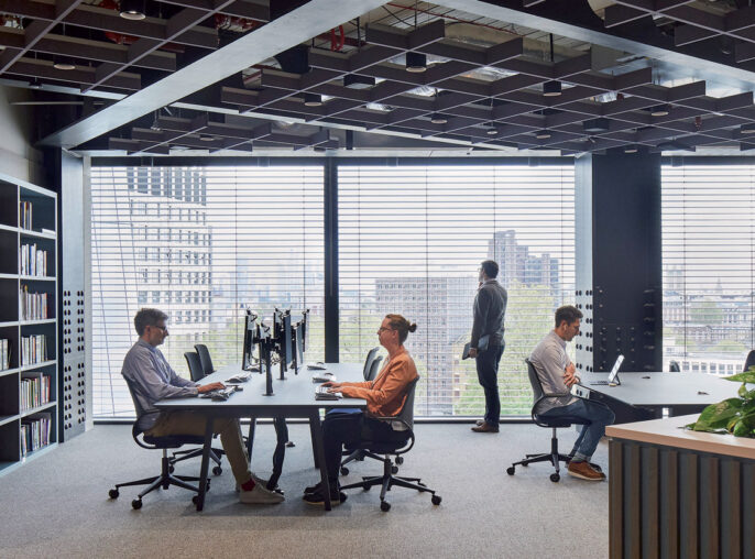 Modern open-plan office space with floor-to-ceiling windows offering cityscape views. Features include a large bookshelf, collaborative workstations, a green wall divider, and exposed ceiling beams, fostering a blend of industrial and biophilic design elements.