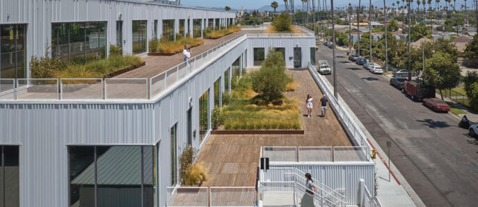 Modern commercial architecture featuring a modular building design with white corrugated metal exteriors. Landscaped green roof terraces and external staircases complement the urban backdrop, enhancing sustainability and communal space.