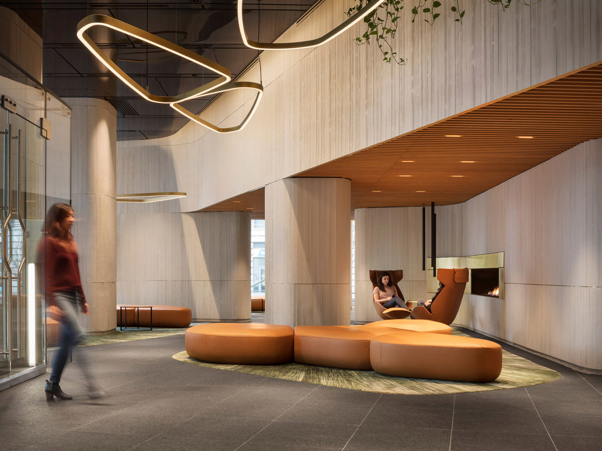 Modern lobby with organic-shaped, tan upholstered seating arrangements centered on a circular green rug, flanked by wooden wall panels and concrete columns. Sinuous, sculptural lighting fixtures hang above, enhancing the contemporary aesthetic. Blurred figures imply motion, suggesting a bustling atmosphere.