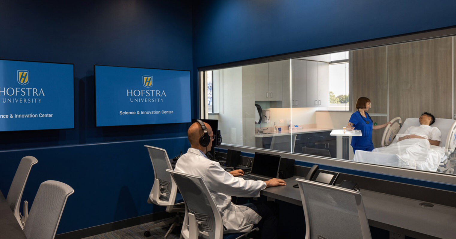 Modern, functional educational facility with observation room featuring ergonomic seating, dual-monitor workstations, glass partition for visibility, and active learning setup with a patient simulation on the adjacent room, showcasing Hofstra University branding on prominently displayed wall signage.