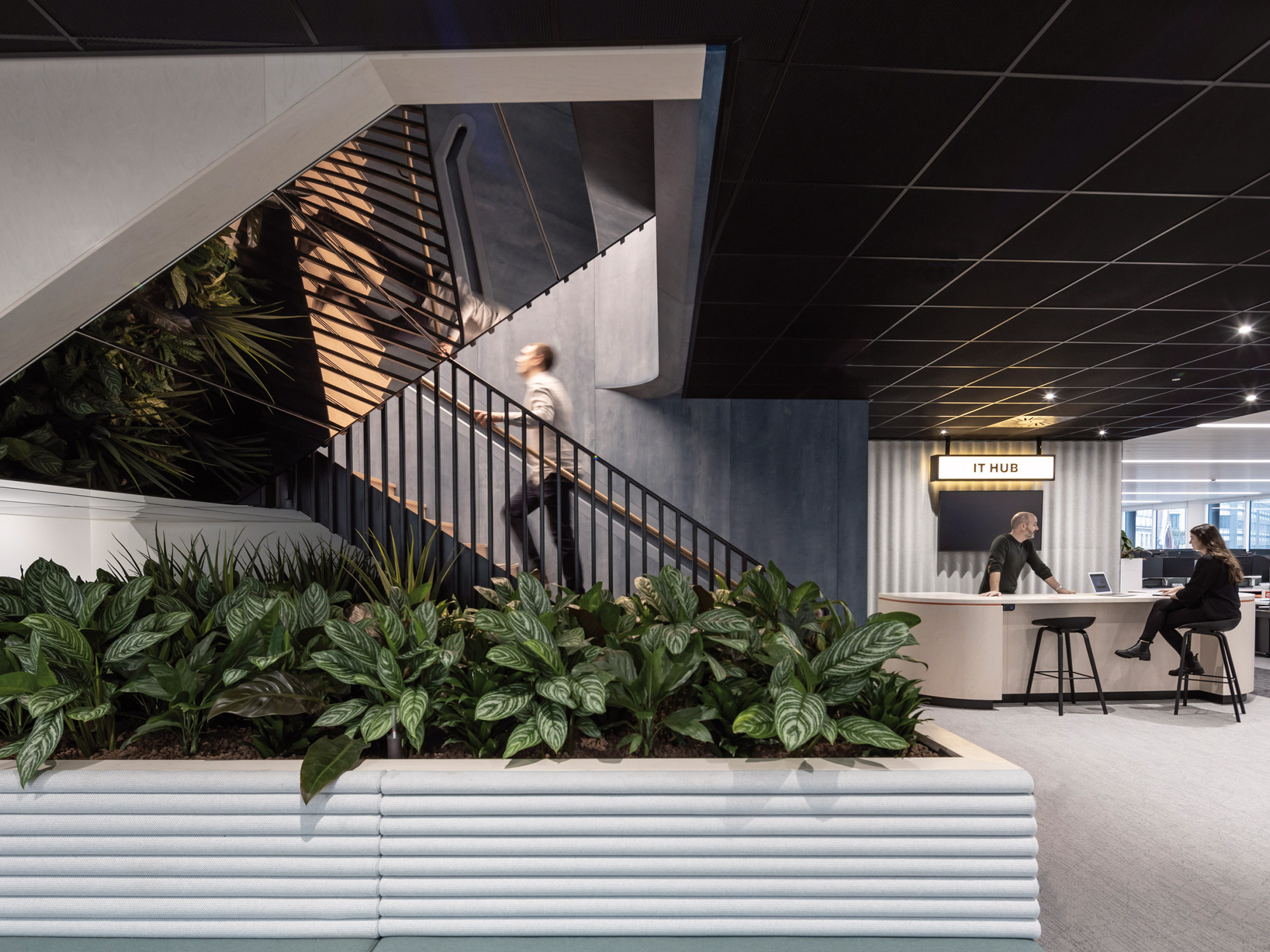 Modern office space featuring a sleek staircase with a person descending, lush green plants at the base, and an it hub counter with an attendant in the background.