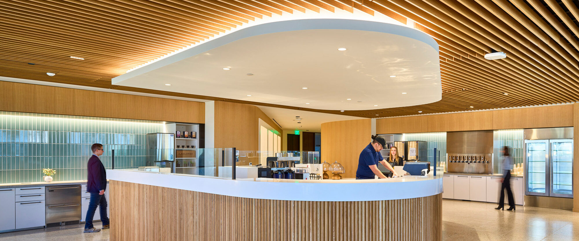 Modern interior showcasing a curved reception desk with vertical wooden slats, complemented by an expansive wooden slatted ceiling that follows the desk's curvature. Neutral tones and ambient lighting enhance the open, airy atmosphere, while individuals interact, suggesting a communal corporate environment.