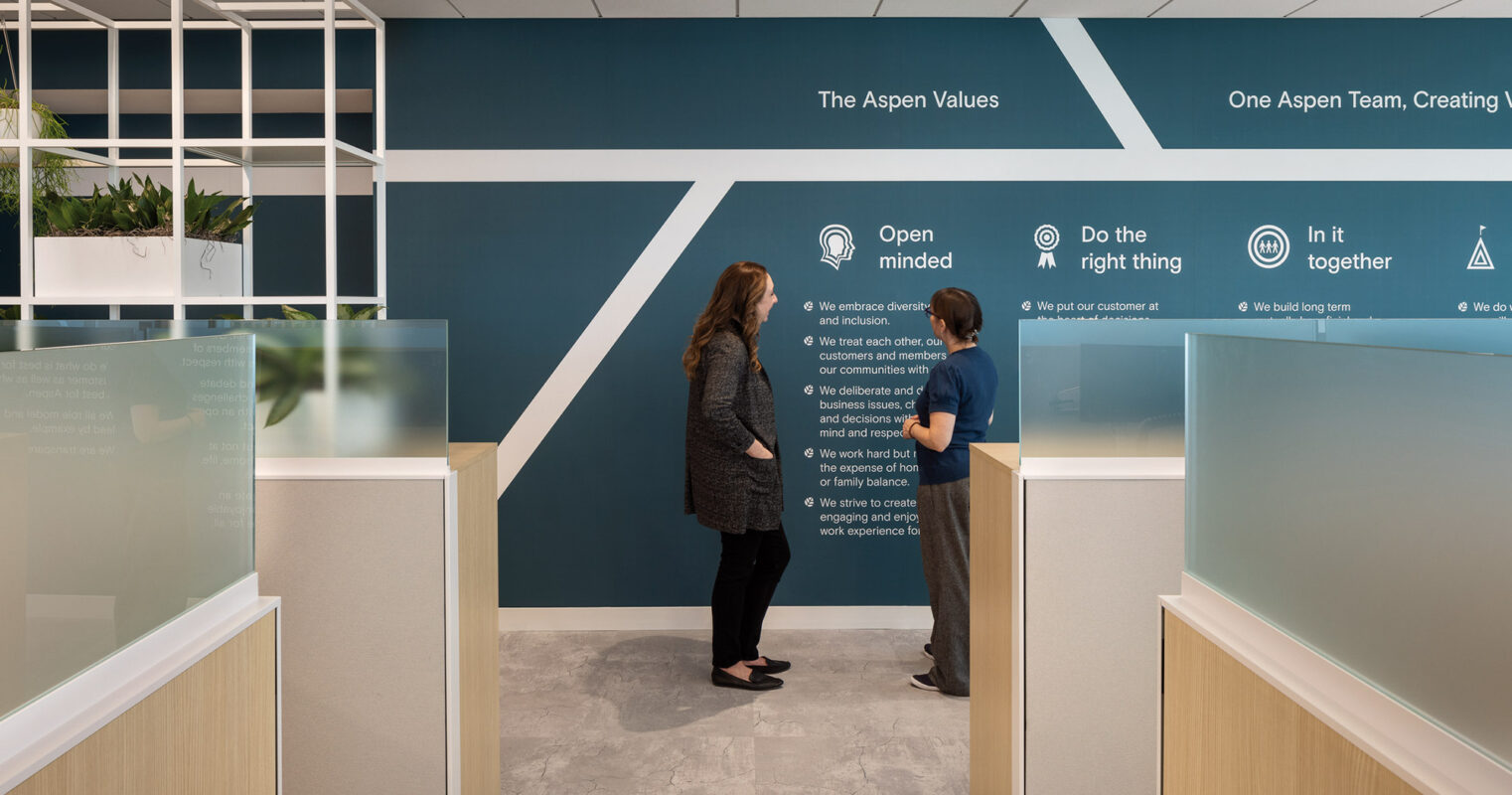 Two individuals in a modern office setting, engaging in conversation beside core value statements displayed on the wall.