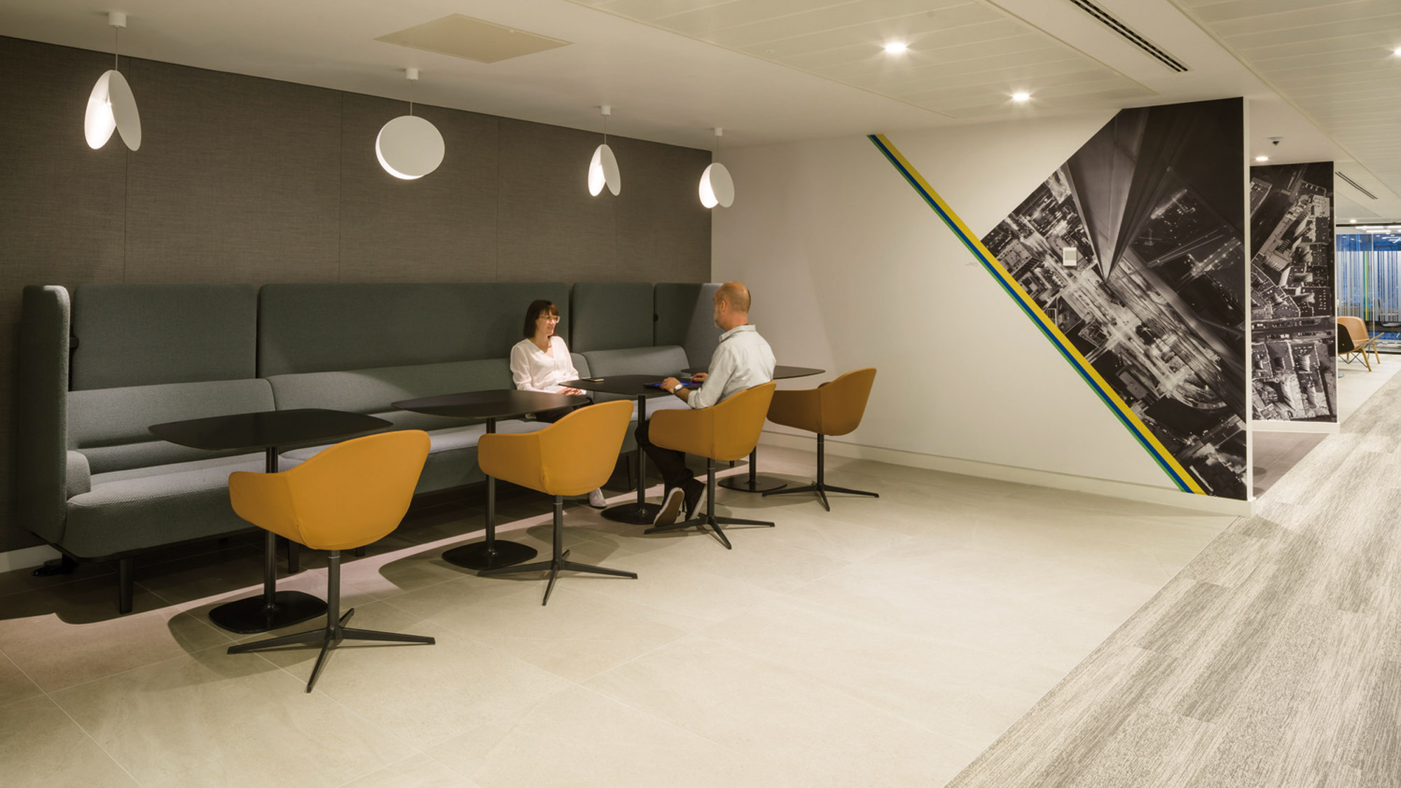 Modern office break area with sleek, gray banquet seating and mustard accent chairs, paired with simple wood-top tables. A monochromatic cityscape mural punctuates the space alongside a bold, diagonally striped accent wall, enhancing the room’s contemporary atmosphere.