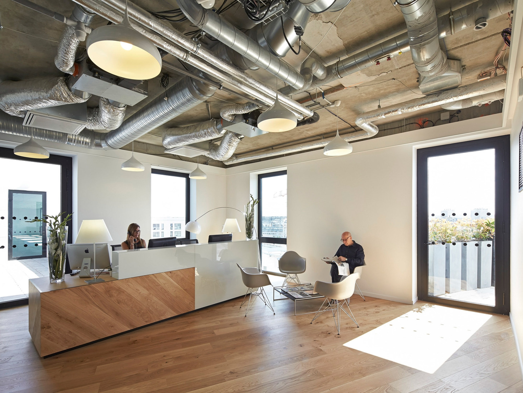 Modern office space featuring exposed ductwork and natural lighting. Varied seating, including ergonomic chairs and lounge areas, complements the light wood floors and white walls, conveying an open, airy atmosphere.