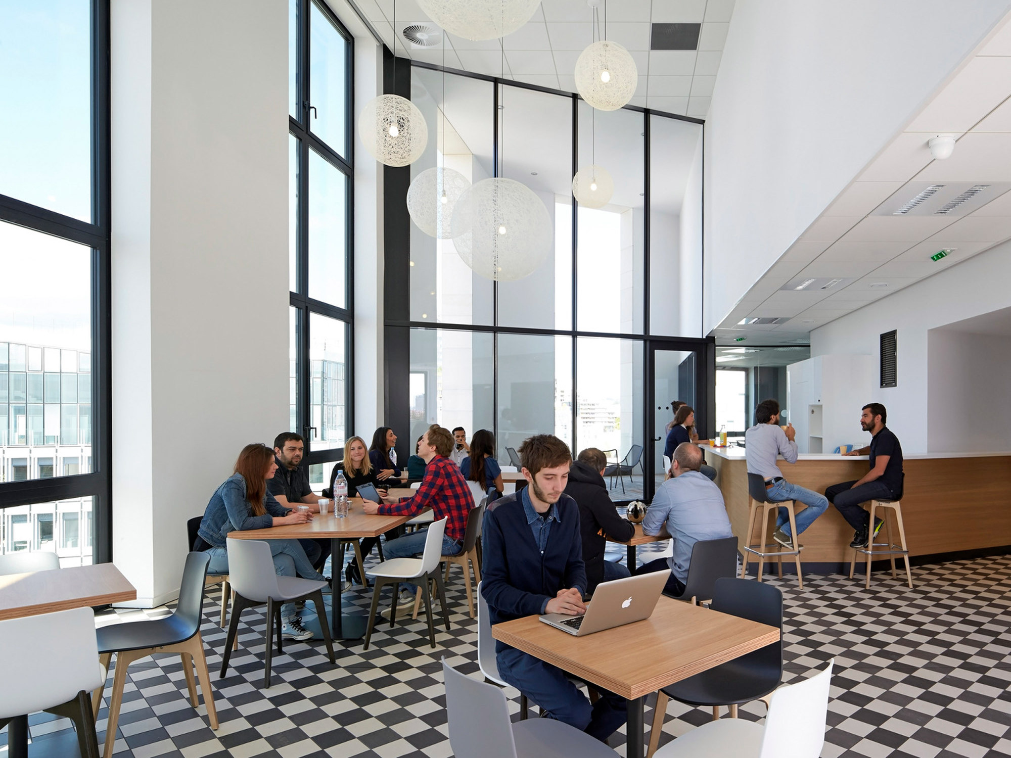 Modern office break room with large windows, geometric patterned flooring, minimalist white pendant lights, and a combination of communal and individual seating areas fostering both collaboration and independent work.