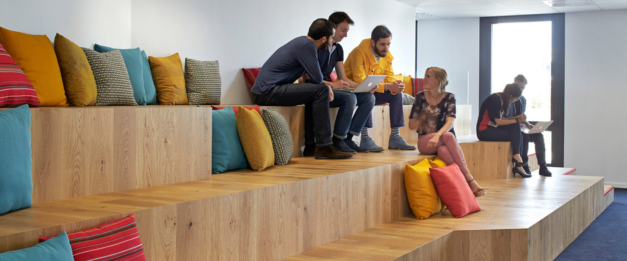 Modern office breakout area with tiered wooden seating steps adorned with colorful cushions. Employees casually engage in conversation, encouraging a collaborative and relaxed workplace atmosphere. Natural light enhances the warm tones of wood, complementing the vibrant fabric hues.