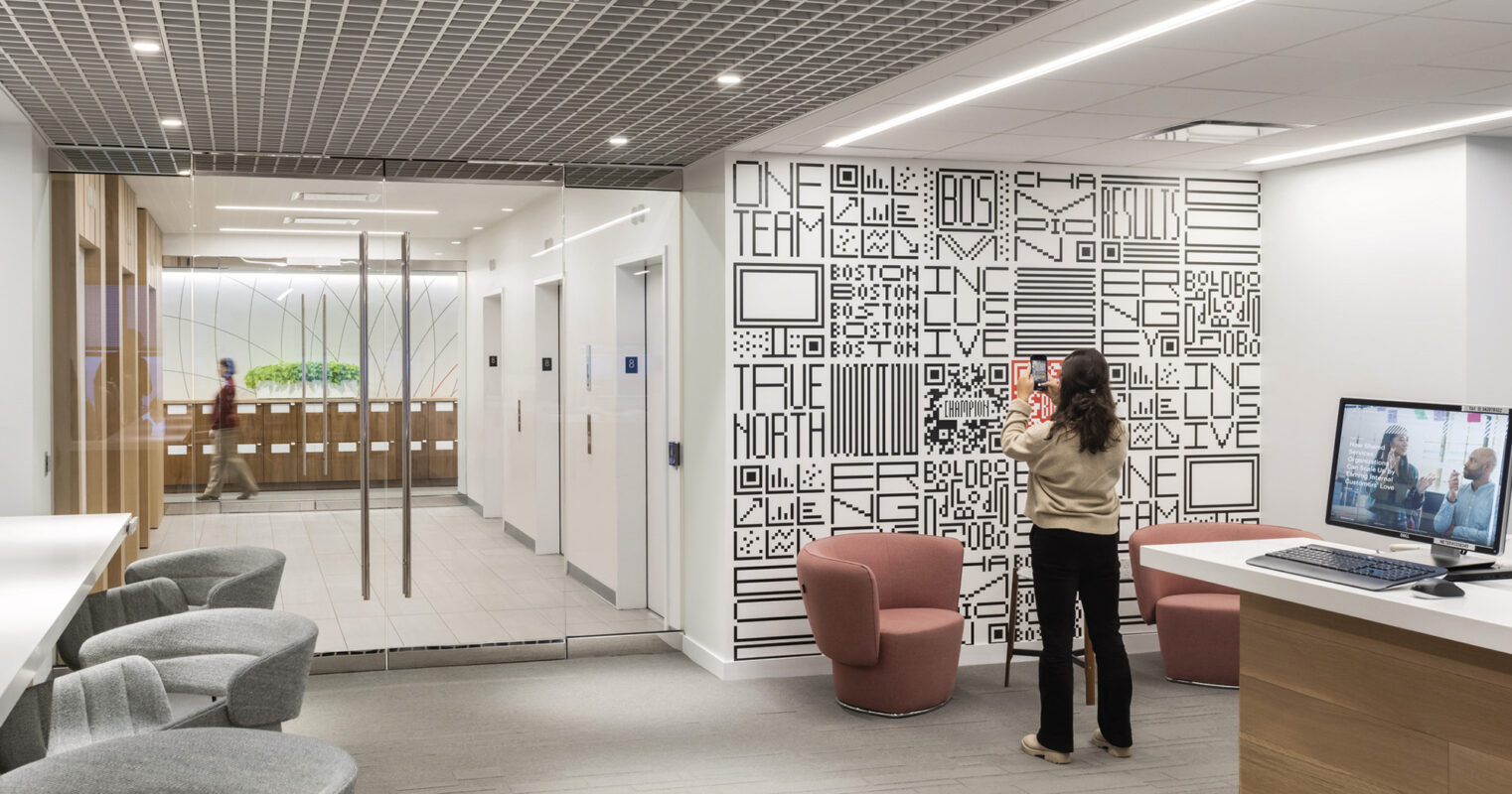 Modern office lobby with stylish interior design, featuring an artistic wall with typographic décor, comfortable seating area, and two individuals engaging with a video conference on a large screen.