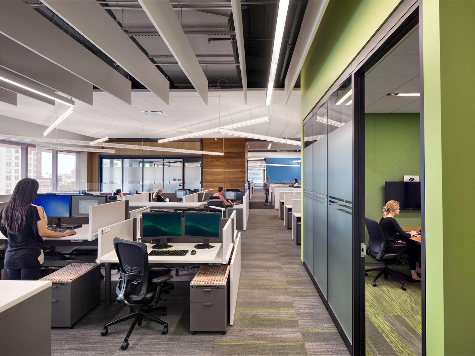 Modern office space with open-floor layout featuring ergonomic workstations, vibrant green glass partitions, subtle gray carpeting, and abundant natural light from floor-to-ceiling windows, complemented by sleek, overhead linear lighting. A professional atmosphere is enhanced by contemporary furniture and unobstructed city views.