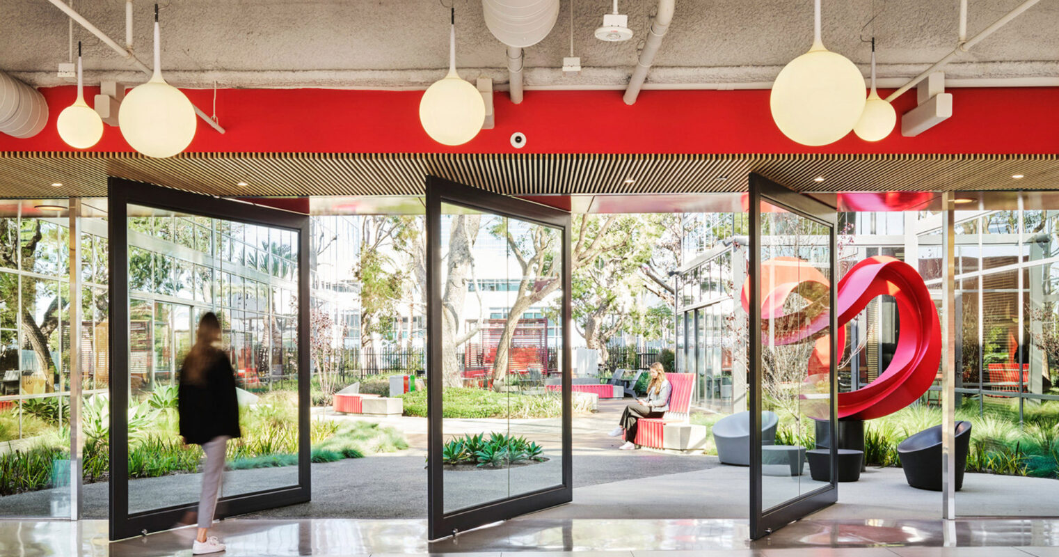 Modern office lobby with floor-to-ceiling windows offering a view of an outdoor green space, accented by a striking red entrance and abstract art installation, complemented by warm wooden slat ceilings and spherical pendant lighting.