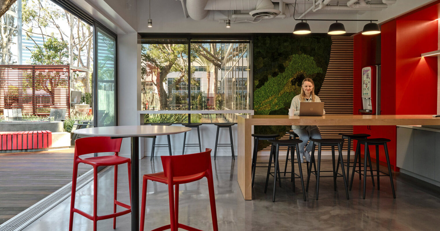 Modern open-plan office with polished concrete floors, featuring a kitchenette with red accents and bar-style seating. Ample natural light filters through floor-to-ceiling windows, complemented by a lush vertical garden and exposed ductwork for an industrial chic aesthetic.