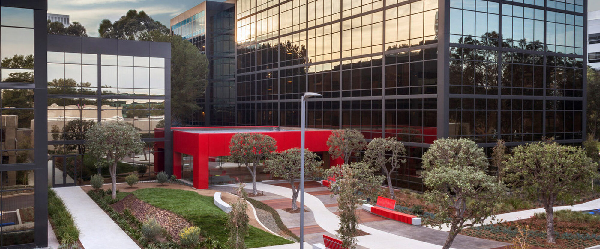 Modern office building exterior featuring geometric glass facade; fronted by vibrant red entrance structure; landscaped with manicured trees and concrete walkways, amidst a backdrop of lush greenery.