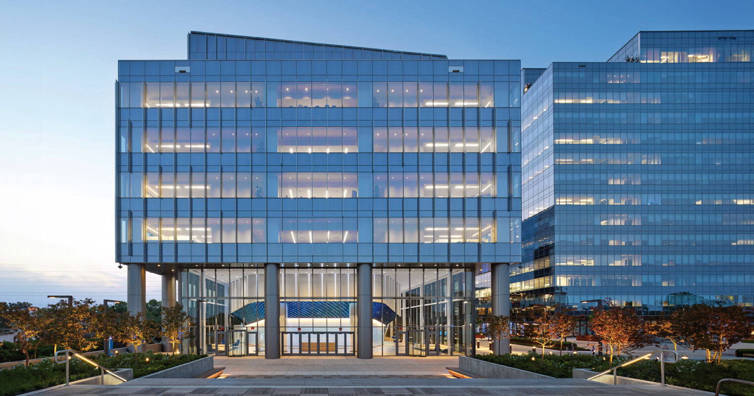 Modern glass office buildings at dusk with illuminated interiors and a calm, clear sky.