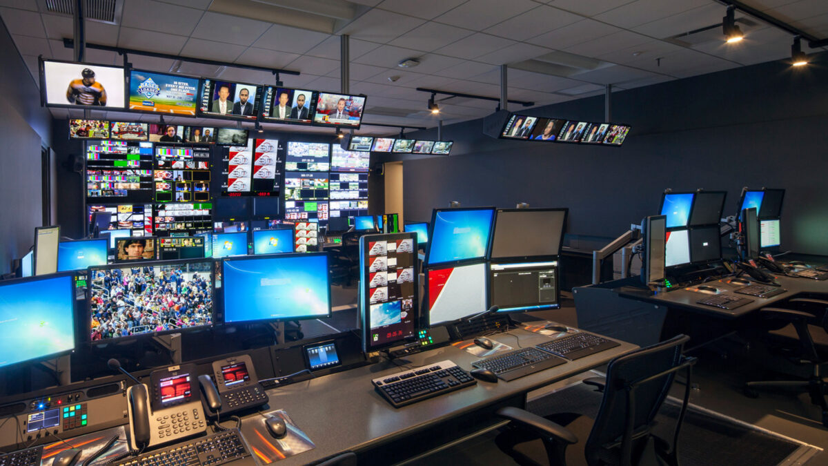 High-tech broadcast control room with multiple screens displaying various channels and content. Sophisticated equipment is arranged on black desks, with ergonomic chairs providing comfort. The space utilizes indirect lighting to minimize glare and enhance concentration for operators.