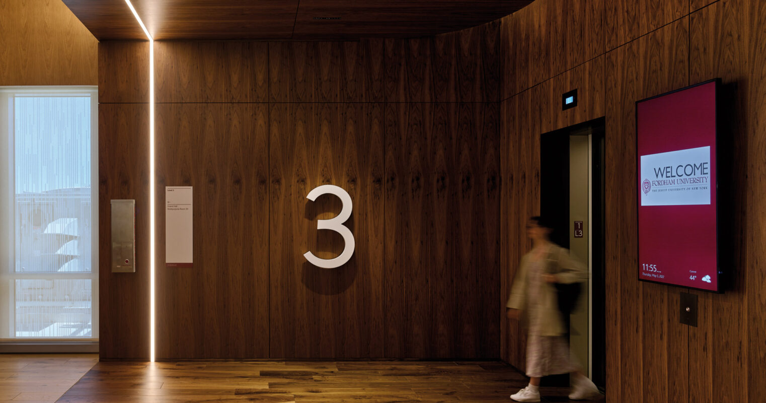 Modern office building interior with a large number '3' on the wall, warm wooden panels, ambient lighting, and a person walking through the hallway.