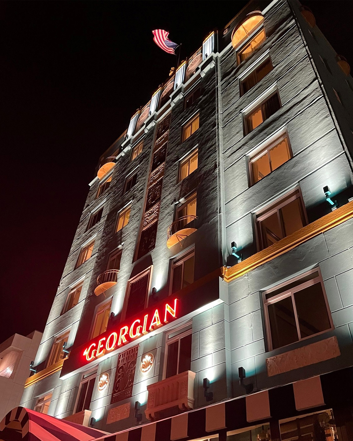 The Georgian hotel illuminated at night with a glowing red neon sign, featuring classic architectural details and an American flag waving atop the building.