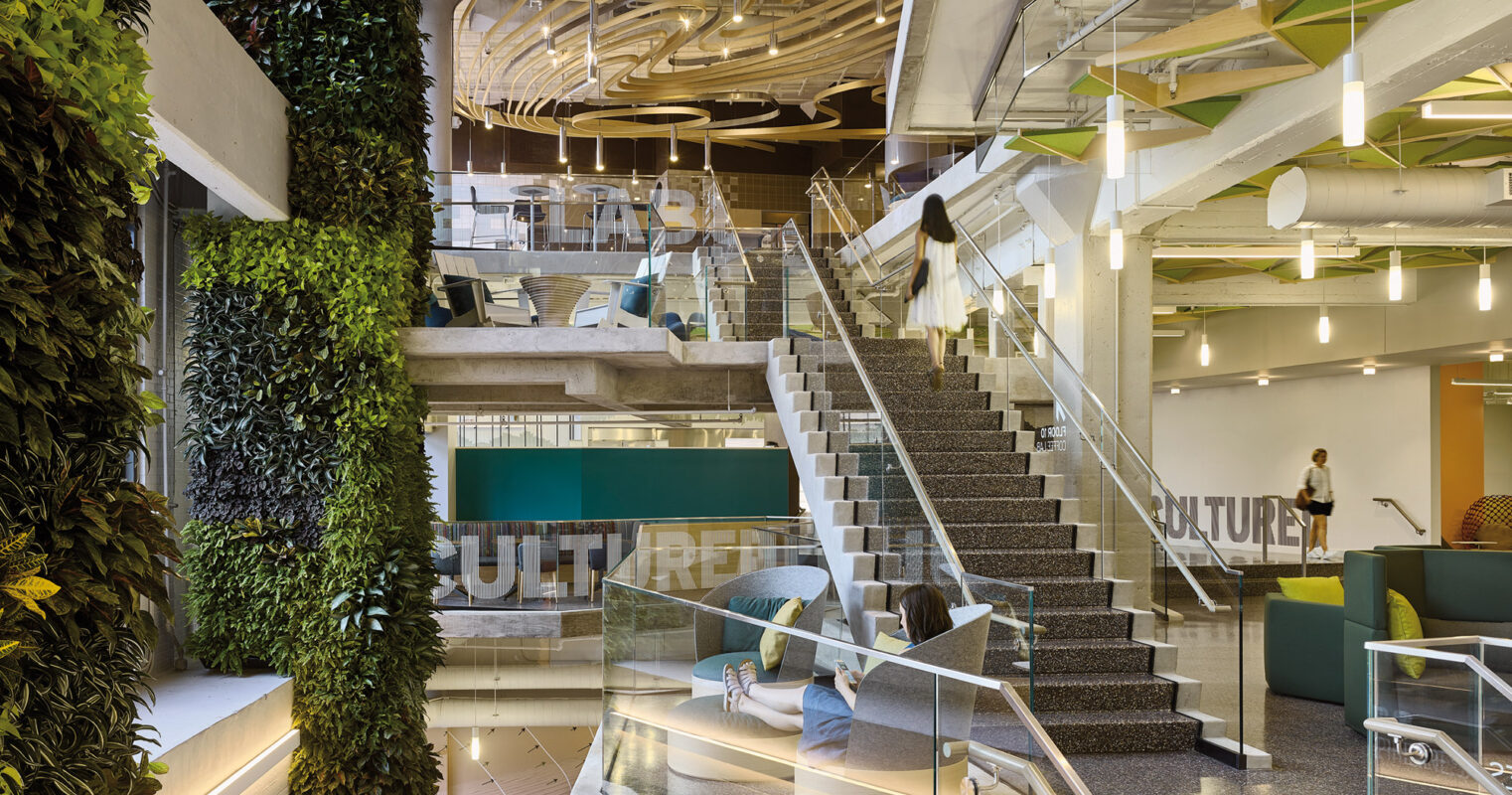 Modern office space with a blend of natural elements, featuring lush green living walls, open staircases, and contemporary design accents, emphasizing sustainability and innovation.