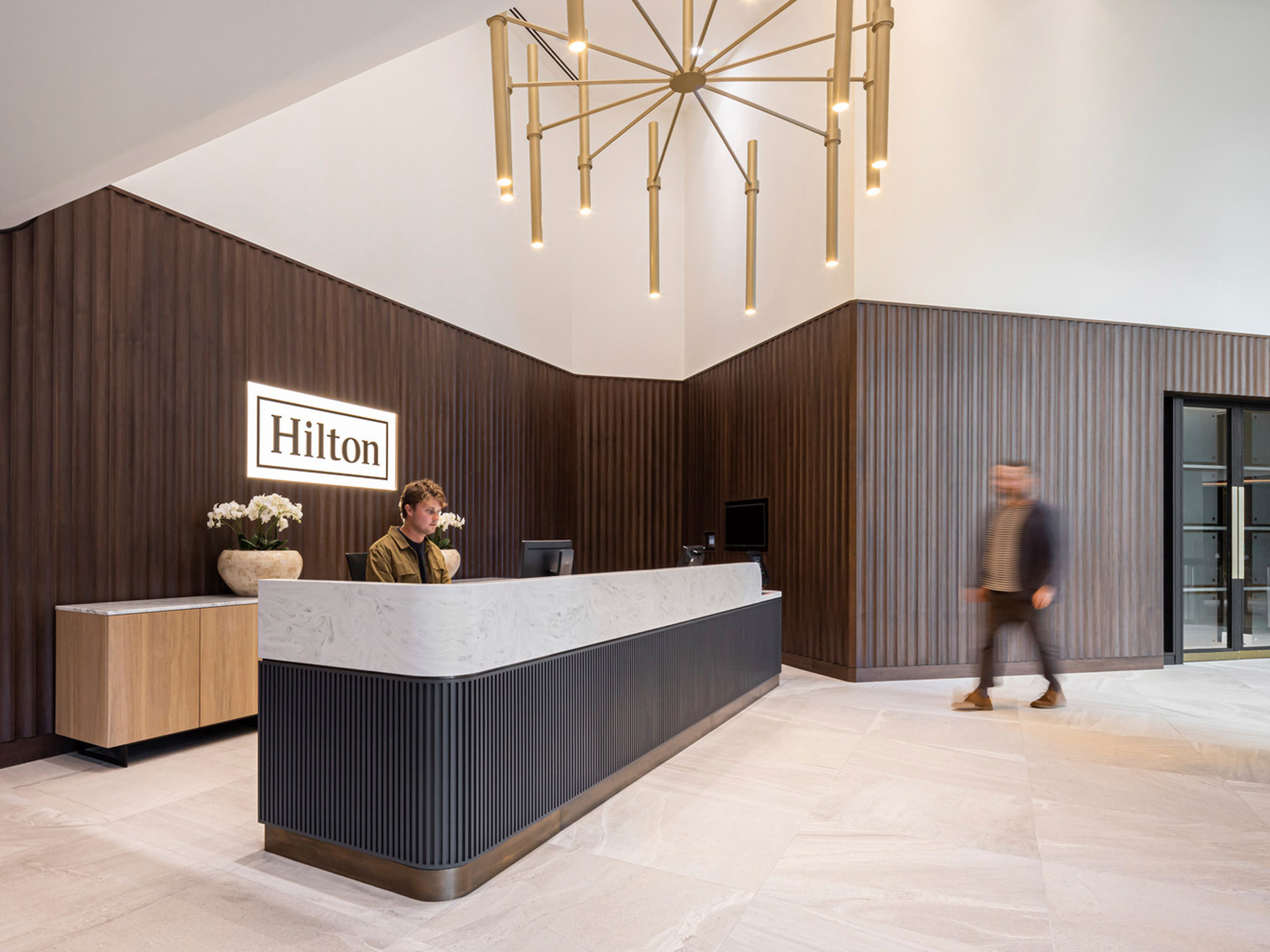 Modern hotel lobby featuring a minimalist walnut reception desk with a white marble top, flanked by cream floor tiles. A geometric gold chandelier hangs above, complementing the sleek, dark wood-panelled walls and ambient lighting. Two individuals provide a sense of scale and activity.