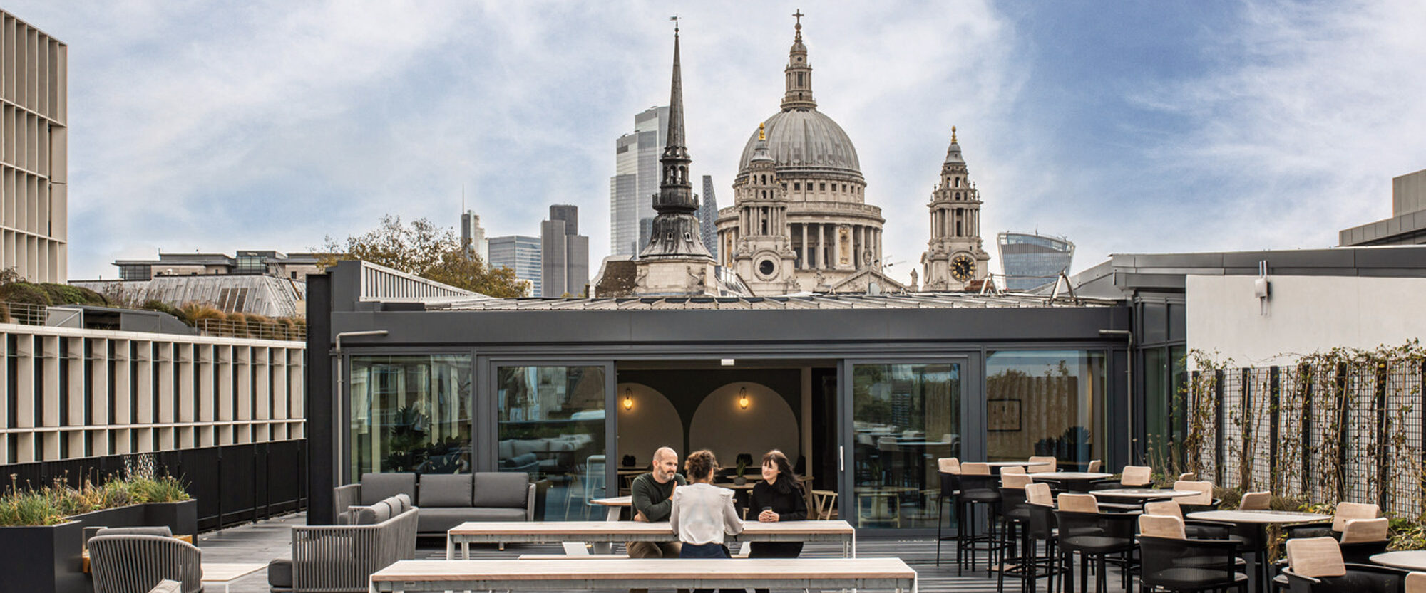 Rooftop terrace with contemporary outdoor furniture, featuring circular dining tables and woven chairs. Lush planters and elegant trellises frame the space, as the backdrop reveals a panoramic view of a city skyline, highlighting the grandeur of St. Paul's Cathedral under a clear sky.