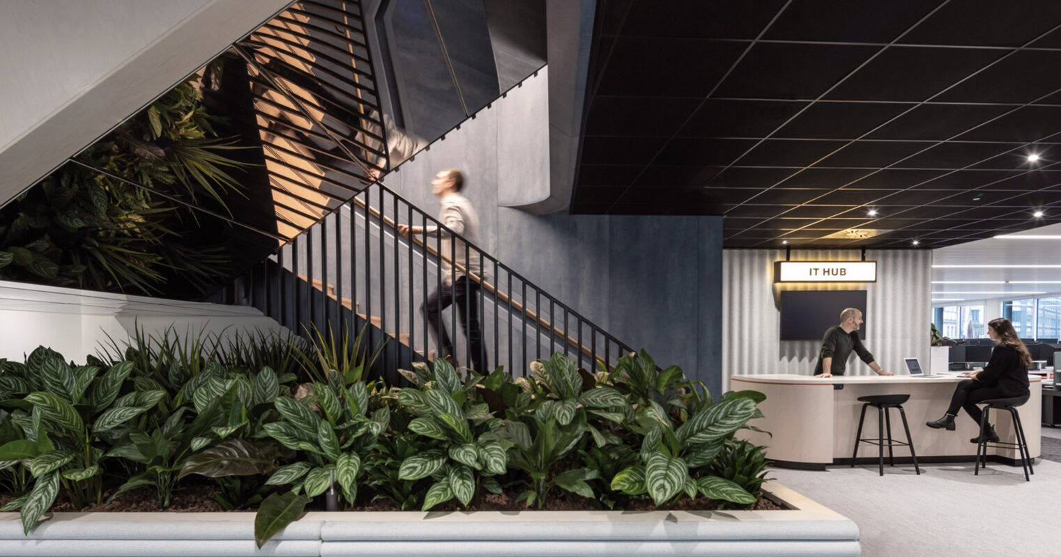 Modern office lobby with a sleek staircase featuring geometric railings, surrounded by lush greenery. The space evokes a dynamic work environment with its contrasting dark ceiling and soft blue upholstered seating area near the reception desk.