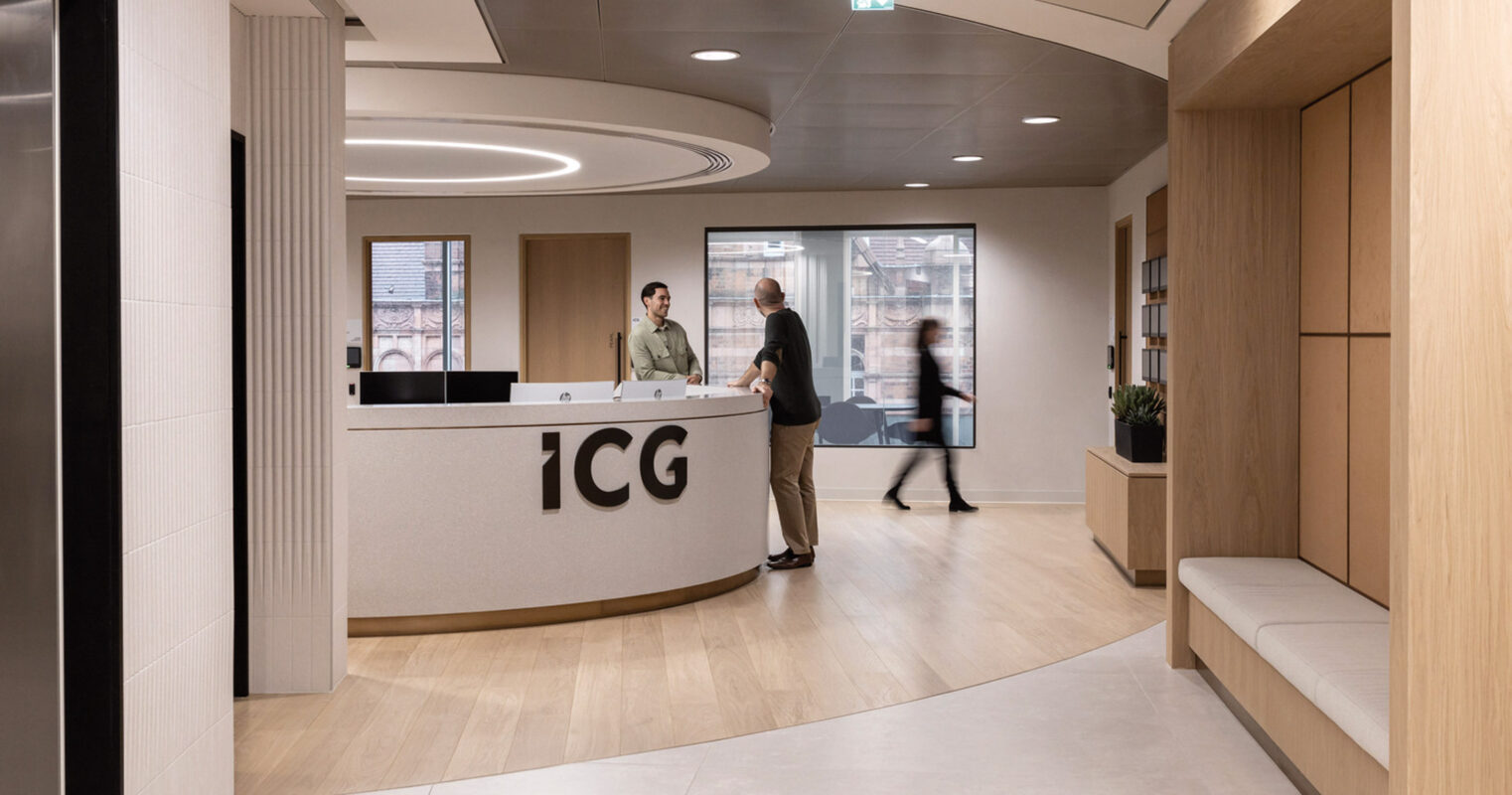 Modern office reception with a sleek, curved desk emblazoned with 'ICG' logo, flanked by warm wood paneling and ambient lighting; visitors converse nearby, clearly navigating a space designed for both function and aesthetics.