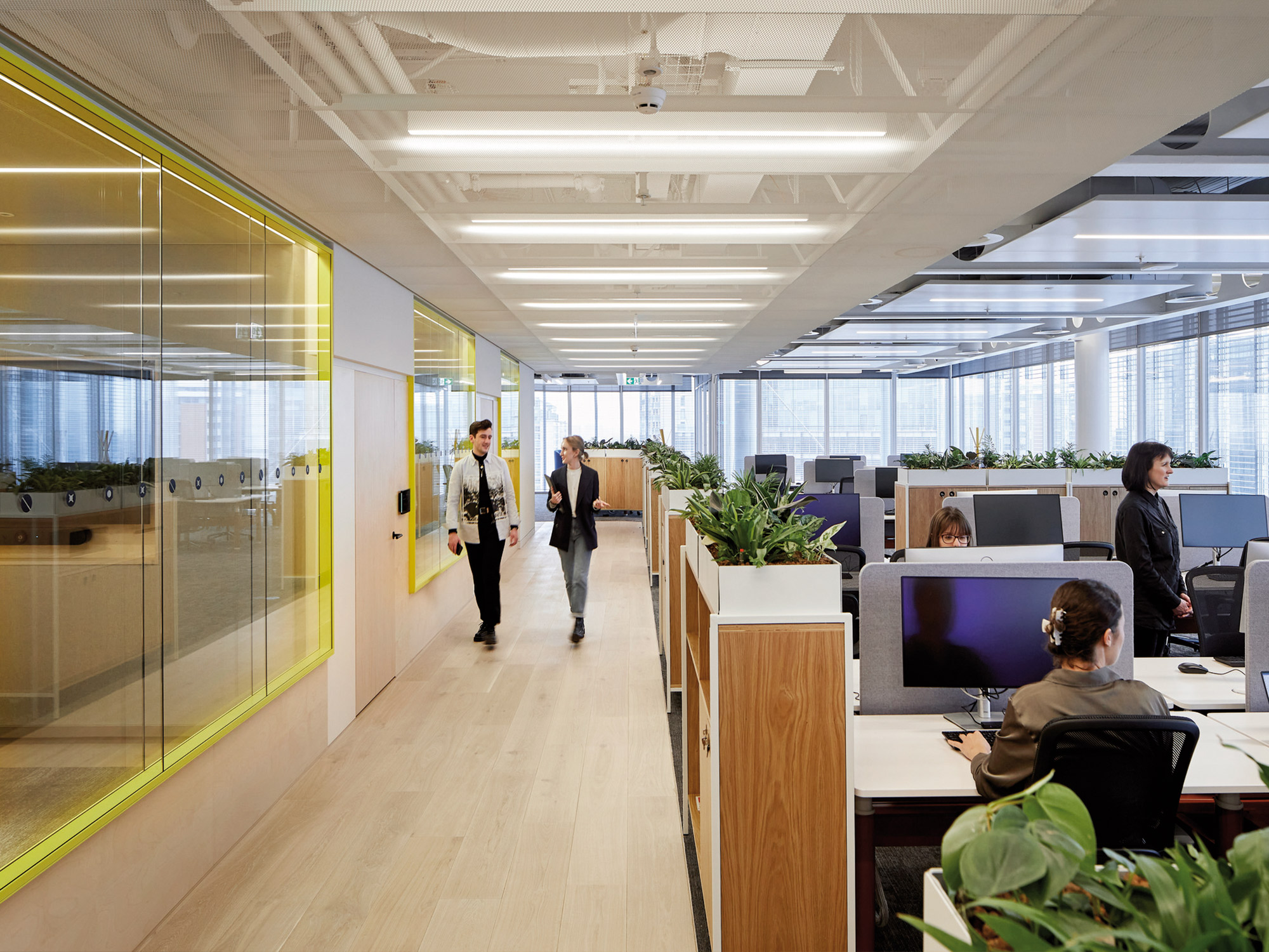 Modern office space bustling with activity, featuring employees engaged in various tasks, with an interior design that incorporates glass partitions, wooden floors, and ample lighting.