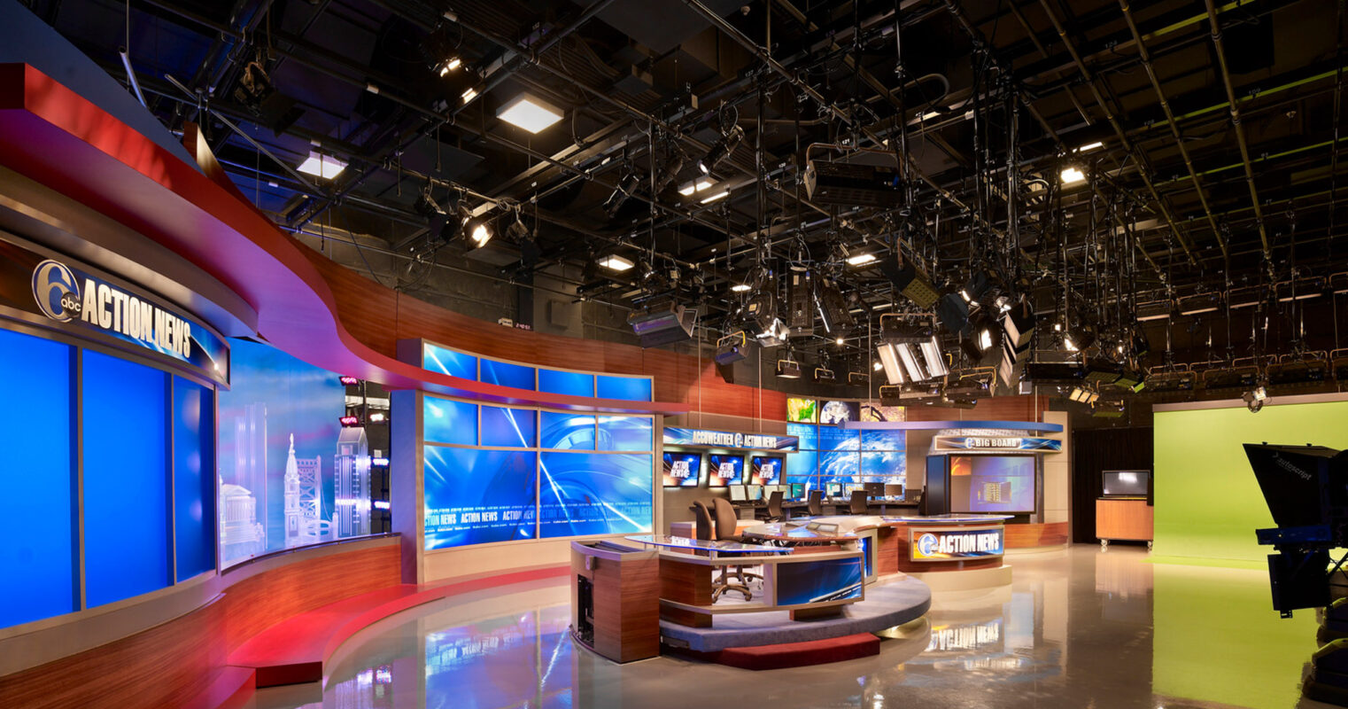 Modern broadcast studio with dynamic lighting and sleek finishes. Central anchor desk with backlit panels, surrounded by high-tech screens and a chroma key green screen for visual effects.